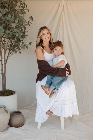 A parent and child pose together on a white chair with a potted eucalyptus plant in the background against a light backdrop.
