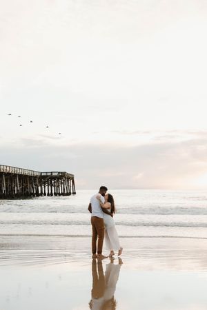 A couple shares a romantic moment at sunset on the beach near a wooden pier.