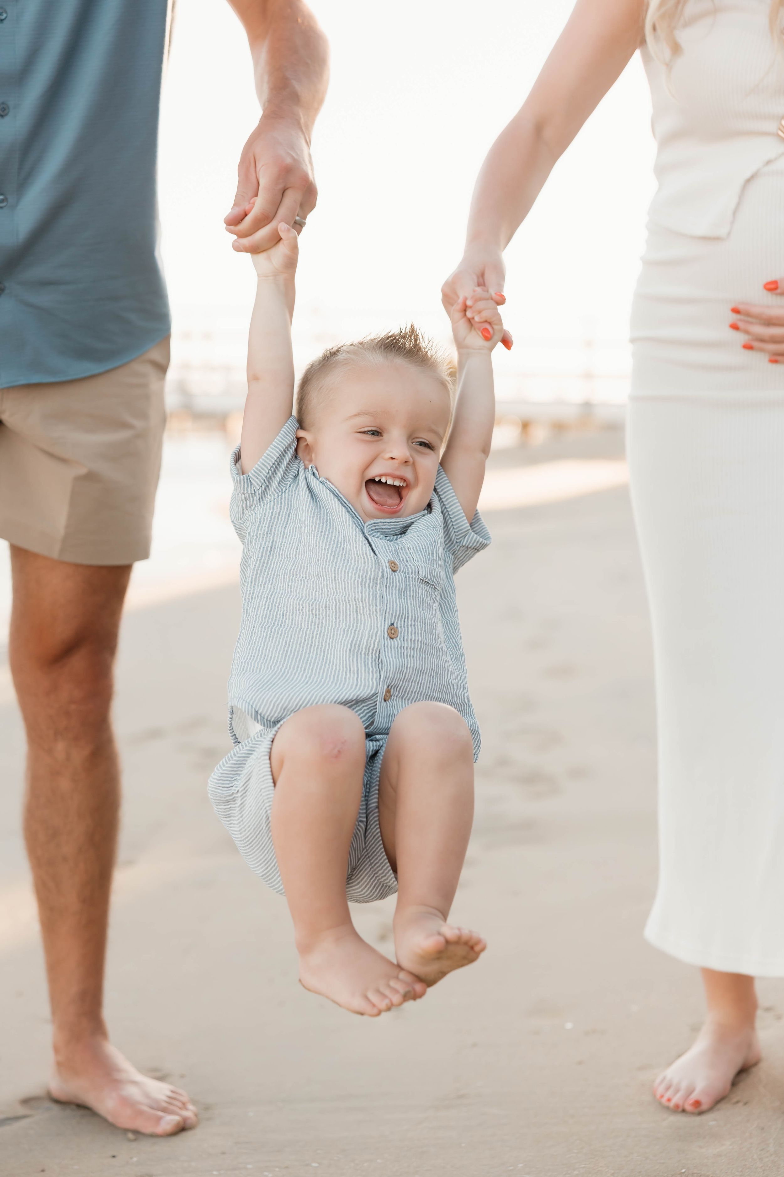Happy toddler boy laughing while being swung by parents hands on a sandy beach in matching blue outfits.