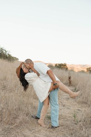 A couple shares a romantic kiss in a golden field, man lifting woman in white dress and tan boots.