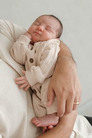 Sleeping newborn baby wearing cream knit cardigan rests peacefully on parent's arm with tiny feet visible.