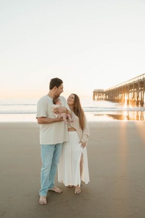 A couple stands barefoot on the beach at sunset with a wooden pier in the background.