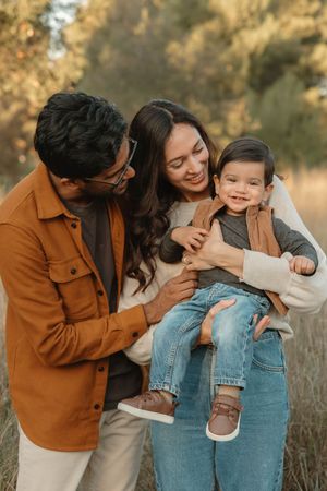 A family of three shares joyful candid moments together in a sunlit outdoor setting wearing casual denim and brown attire.