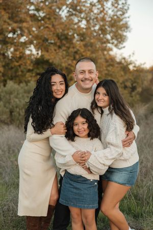 A family wearing coordinated cream and white outfits embraces while laughing together in an outdoor autumn setting.