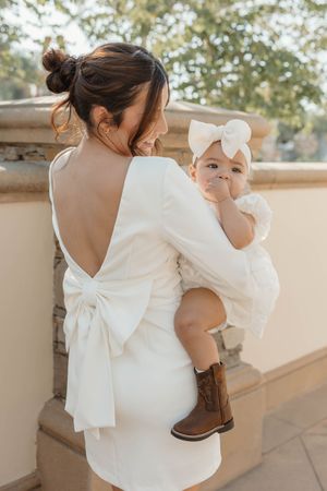 A mother in a white dress with a large bow detail holds her baby while standing on a porch in a photo series.
