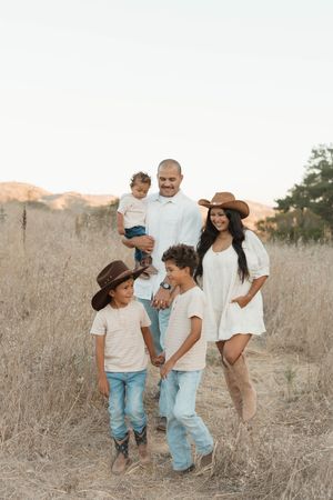 Family of five posing in golden field, parents in white outfits, kids in cowboy hats and jeans, warm sunset lighting.