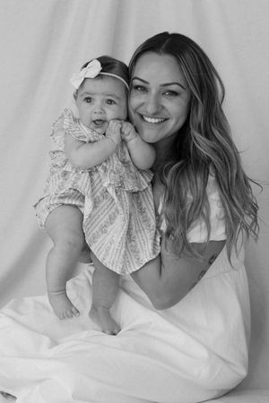 Black and white portrait of a parent and baby in white dresses sitting together against a light backdrop.