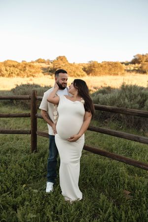 Romantic couple embraces in white dress and casual wear against rustic wooden fence during golden hour sunset.