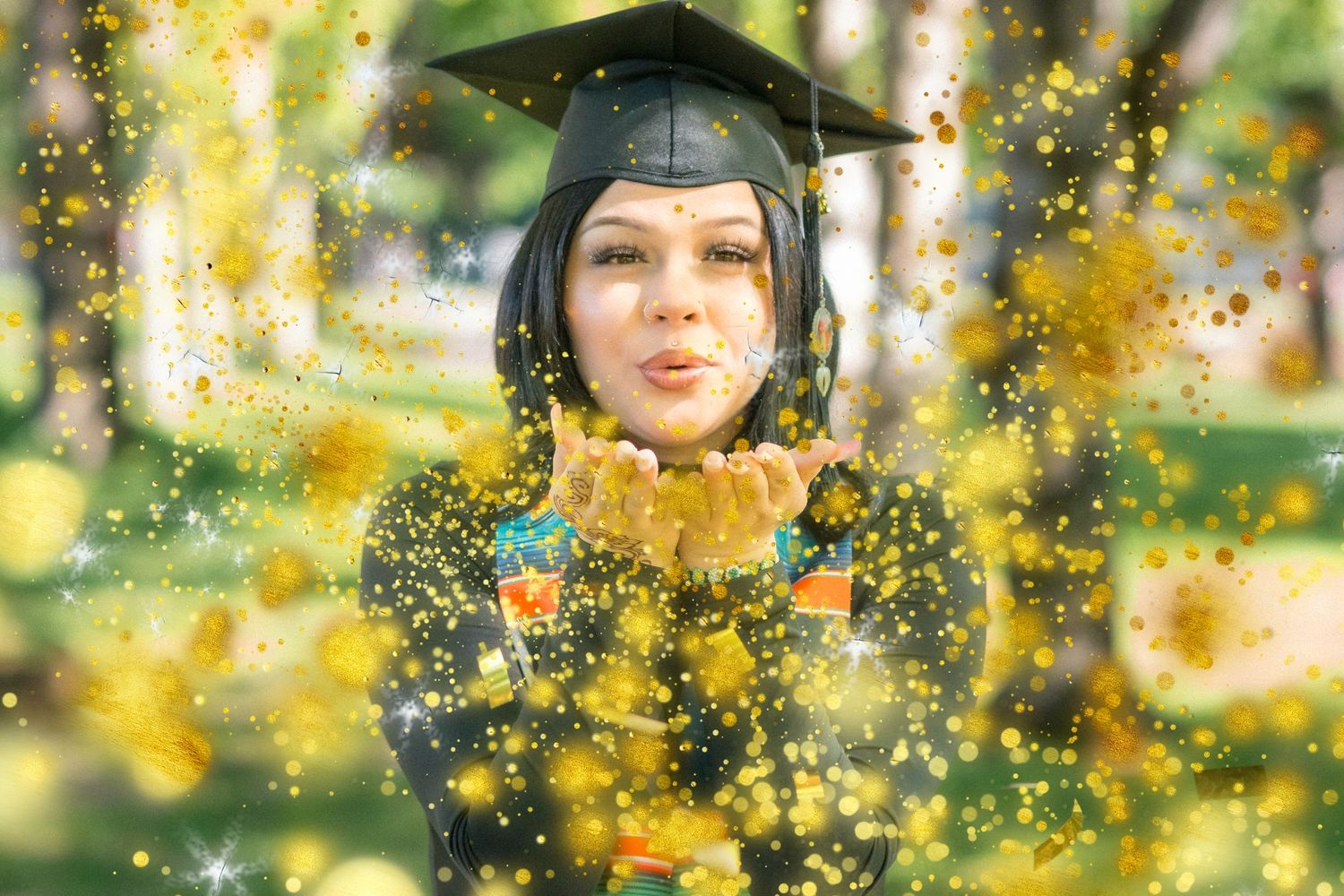 Graduate in cap and gown surrounded by golden sparkles in outdoor setting.