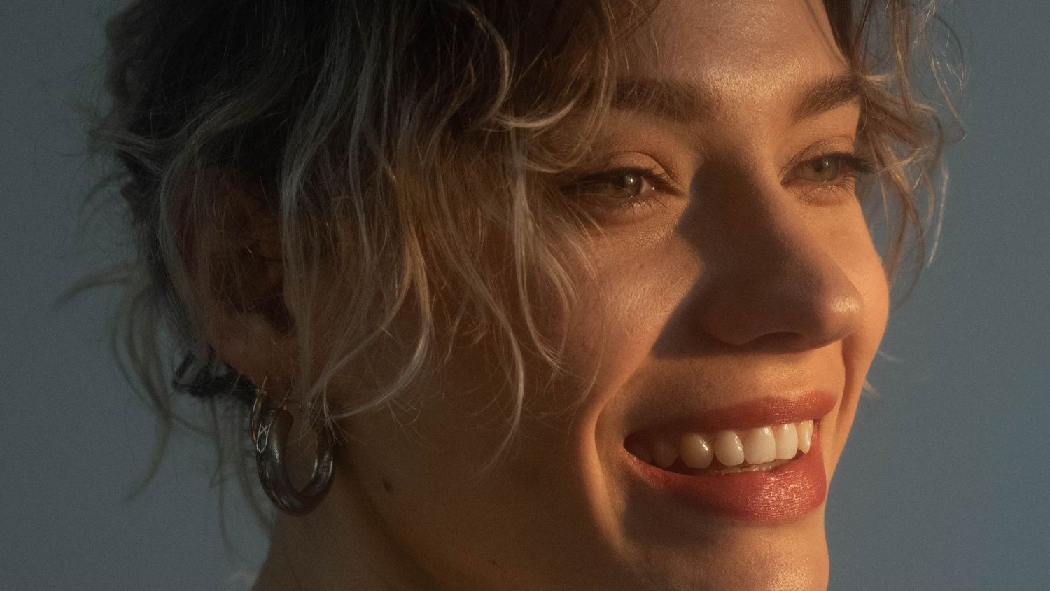 Portrait of a person with curly hair smiling warmly in golden sunlight.