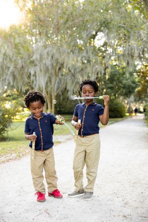 Two children in matching navy shirts and khaki pants play and dance together on a white gravel path surrounded by trees.