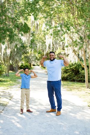 Two people in light blue shirts strike playful poses on a tree-lined path with Spanish moss overhead.
