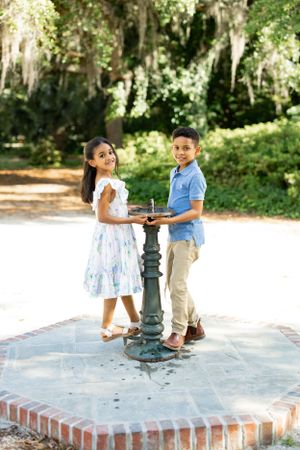 Two young people pose together at a decorative water fountain in a park setting.