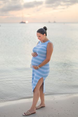 Side view of expectant mother in blue dress walking on beach at dusk.