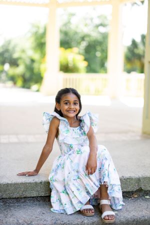 A young person in a floral dress sits outdoors smiling brightly.