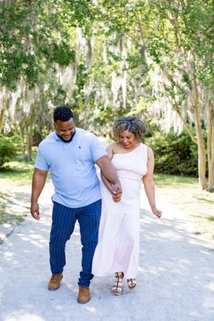 A couple in coordinating blue and pink outfits walks along a scenic path with hanging moss.