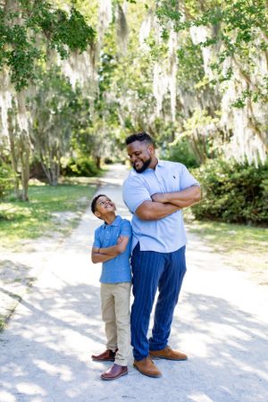 Parent and child strike matching poses with crossed arms on a tree-lined path.