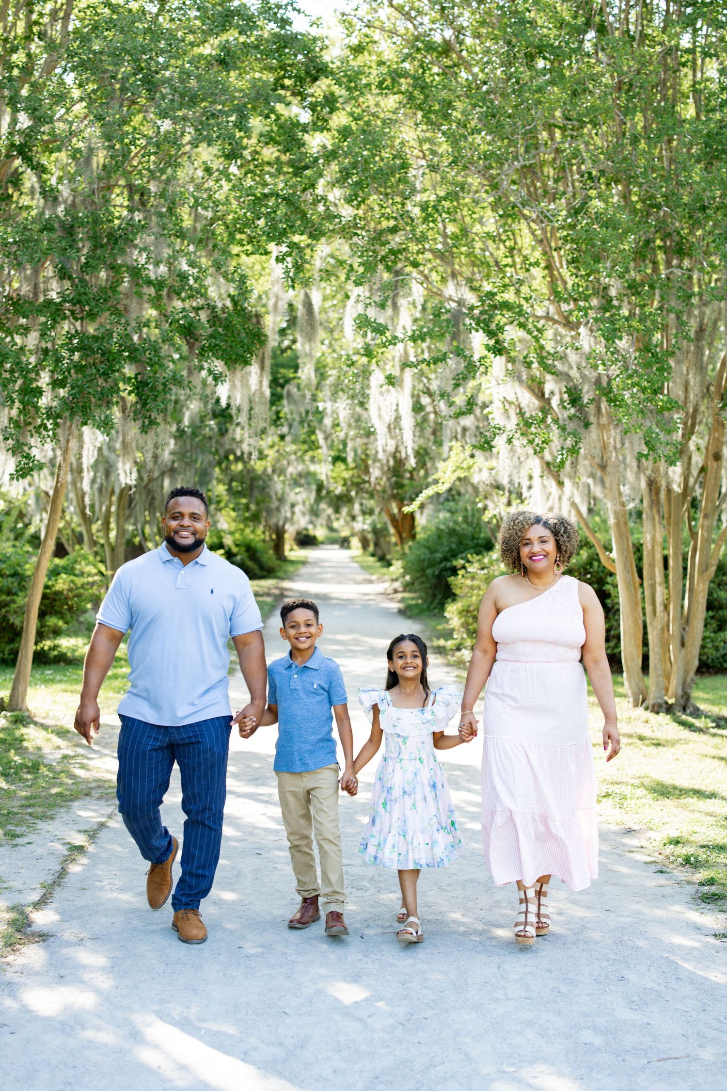 A family of four walking together hand in hand down a tree-lined path on a sunny day.