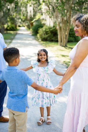A family holds hands in a circle on a sunlit path surrounded by trees.