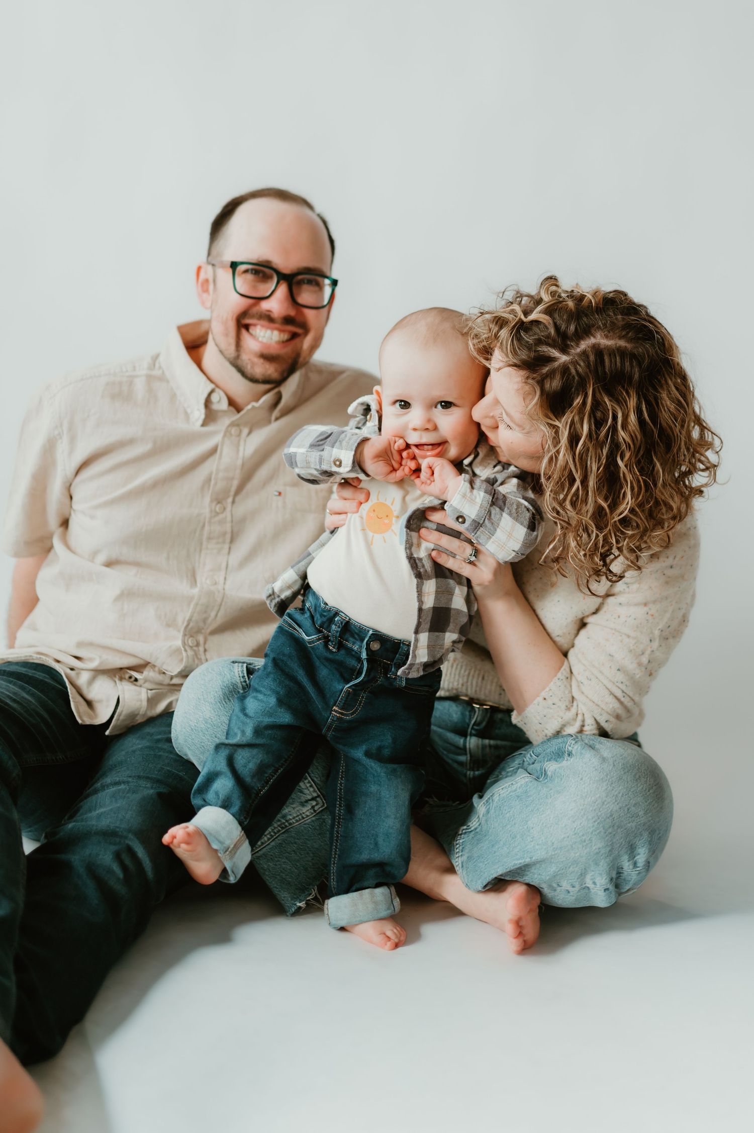 A smiling family with a man and woman kissing a baby in plaid shirt and jeans on white background during family session in Milwaukee Studio
