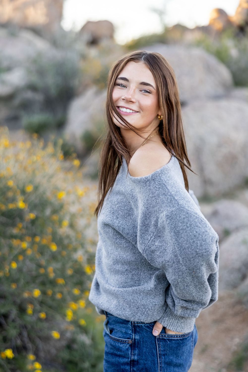 Girl in gray sweater and jeans poses against natural backdrop with yellow wildflowers.
