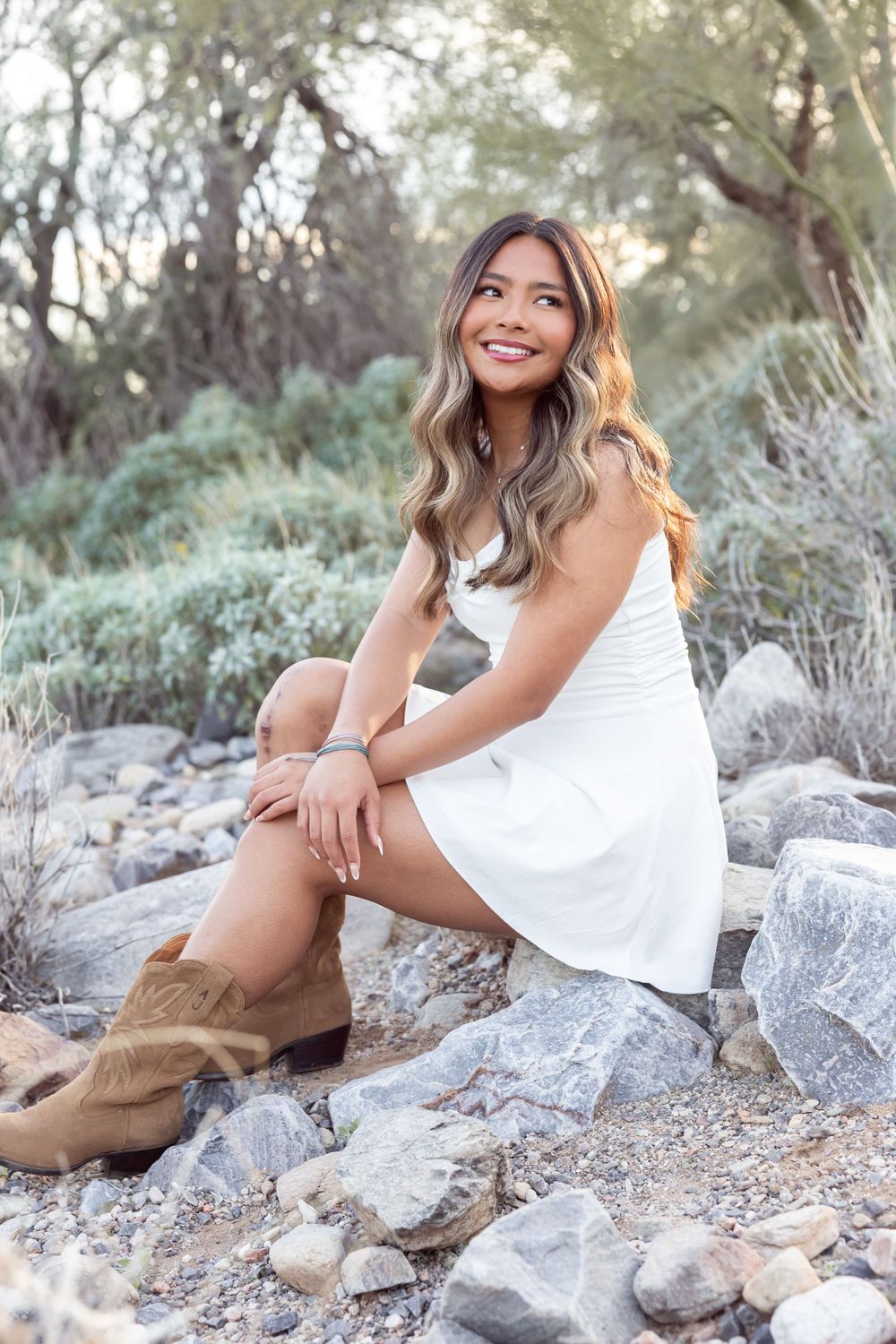 Woman in flowing white dress sits elegantly on rocky terrain during sunset photoshoot.