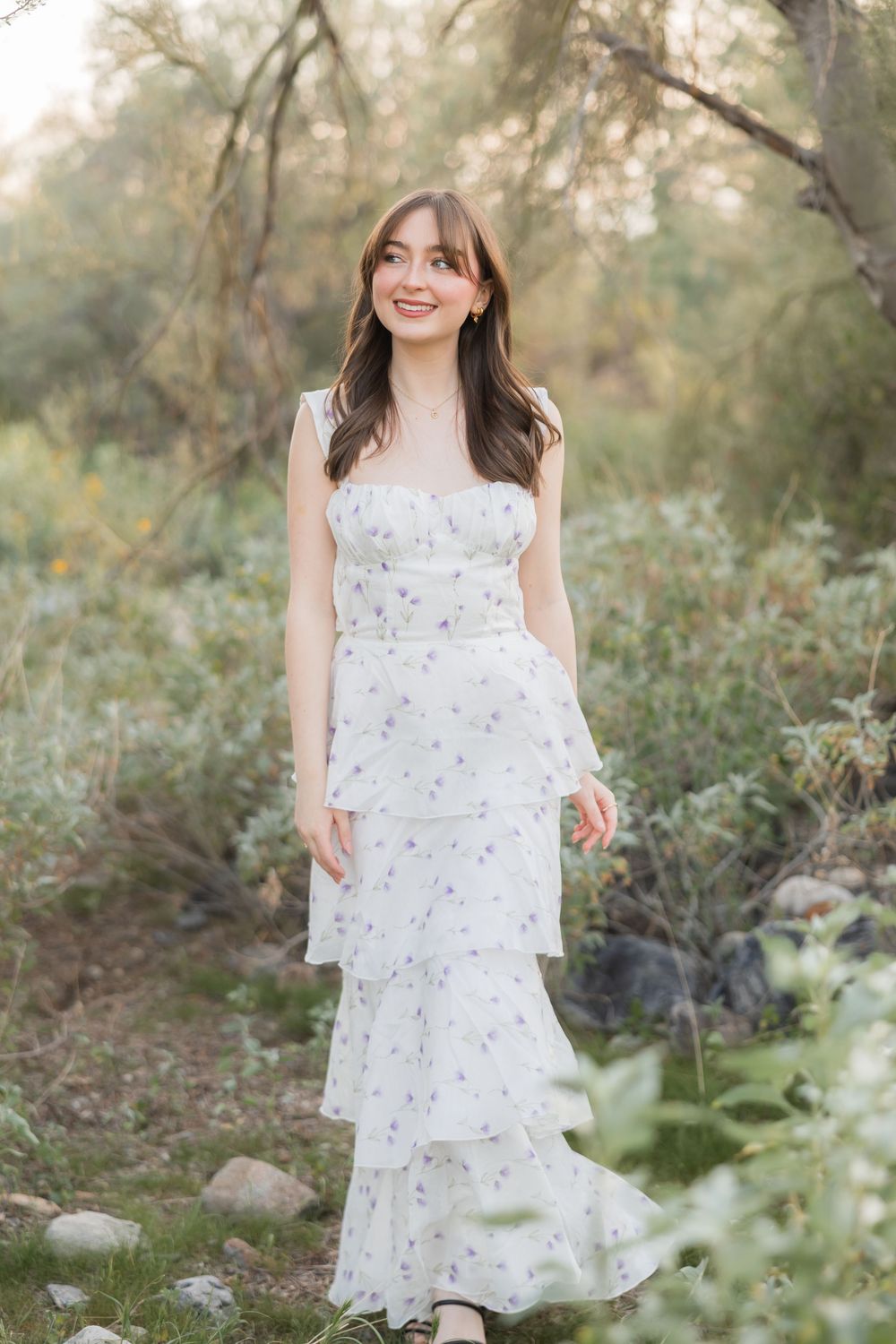 A white floral dress is photographed in a natural outdoor setting with soft lighting and greenery.