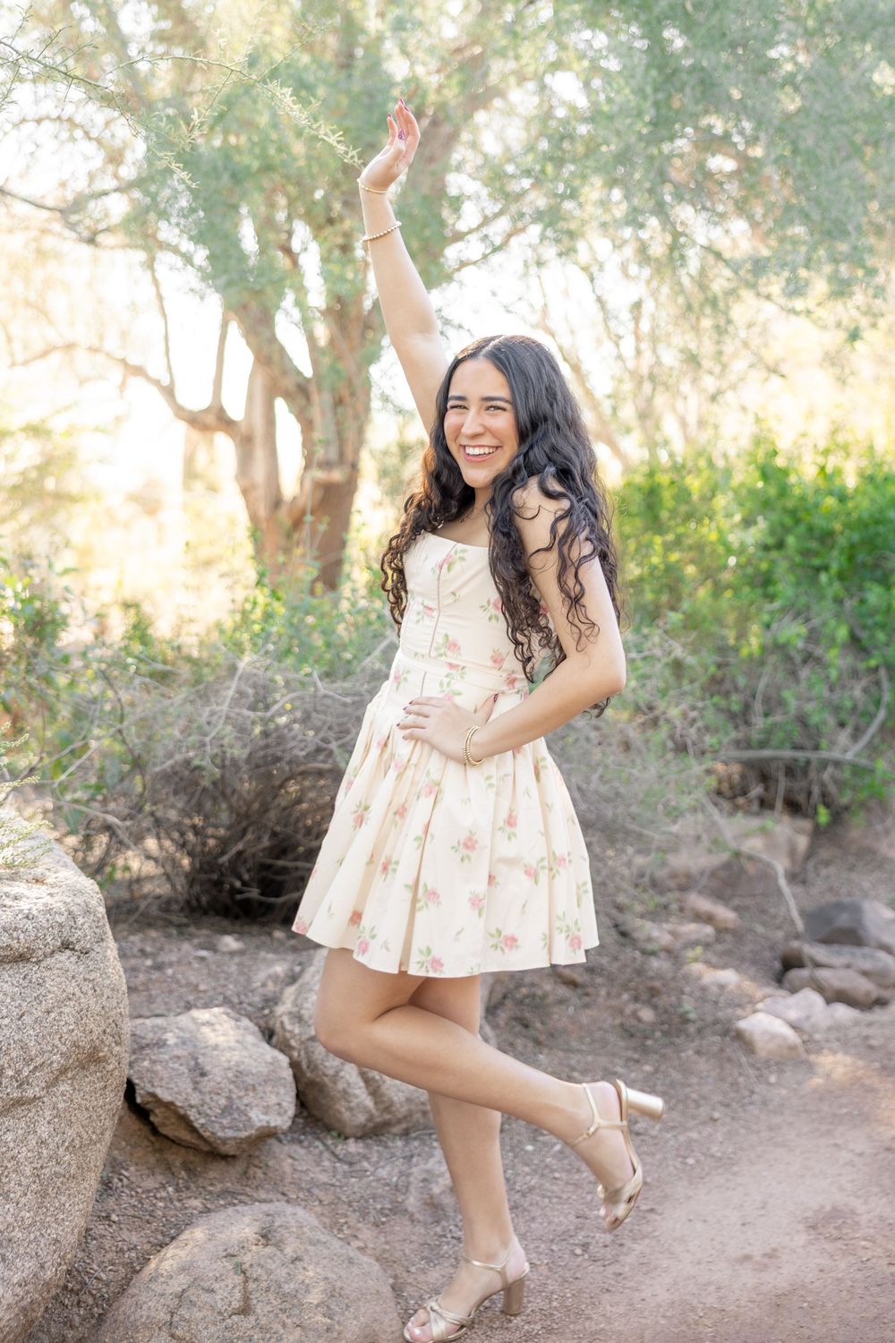 Young woman in floral dress dances playfully in natural outdoor setting with rocks and trees.
