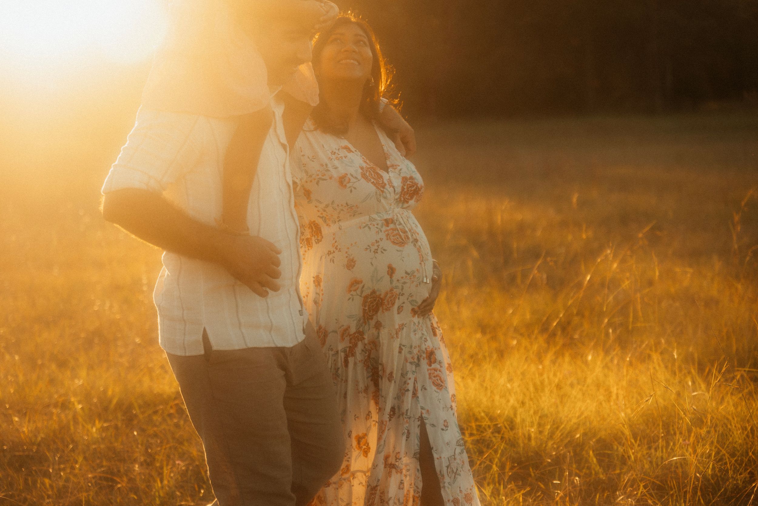 A pregnant figure stands in a golden sunlit field wearing a flowing lace dress at sunset.
