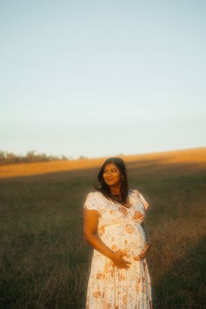 A woman in a floral dress cradles her pregnant belly while standing in a sunlit field.