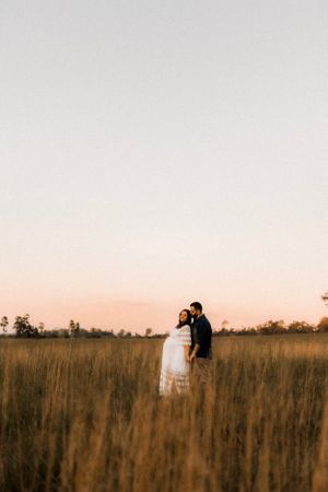 A couple stands close together in tall grass during sunset.