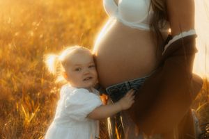 A small child in white clothing embraces a pregnant person's belly in a sunlit field at golden hour.