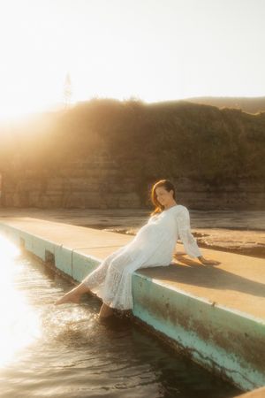 A person in a white dress sits on a seaside wall during golden hour sunset, creating a dreamy coastal atmosphere.