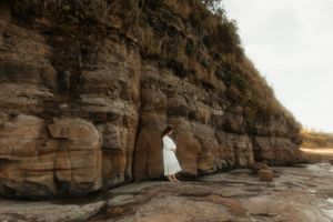 A figure in white stands in a natural rock arch formation along a coastal cliff at sunset.