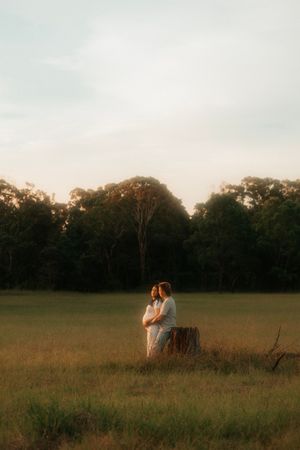 A couple embraces in a rustic field at golden hour with forest treeline silhouetted against the dusky sky.