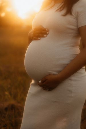 A silhouetted pregnant woman in a white dress holds her belly during sunset, creating a warm and glowing maternity portrait.