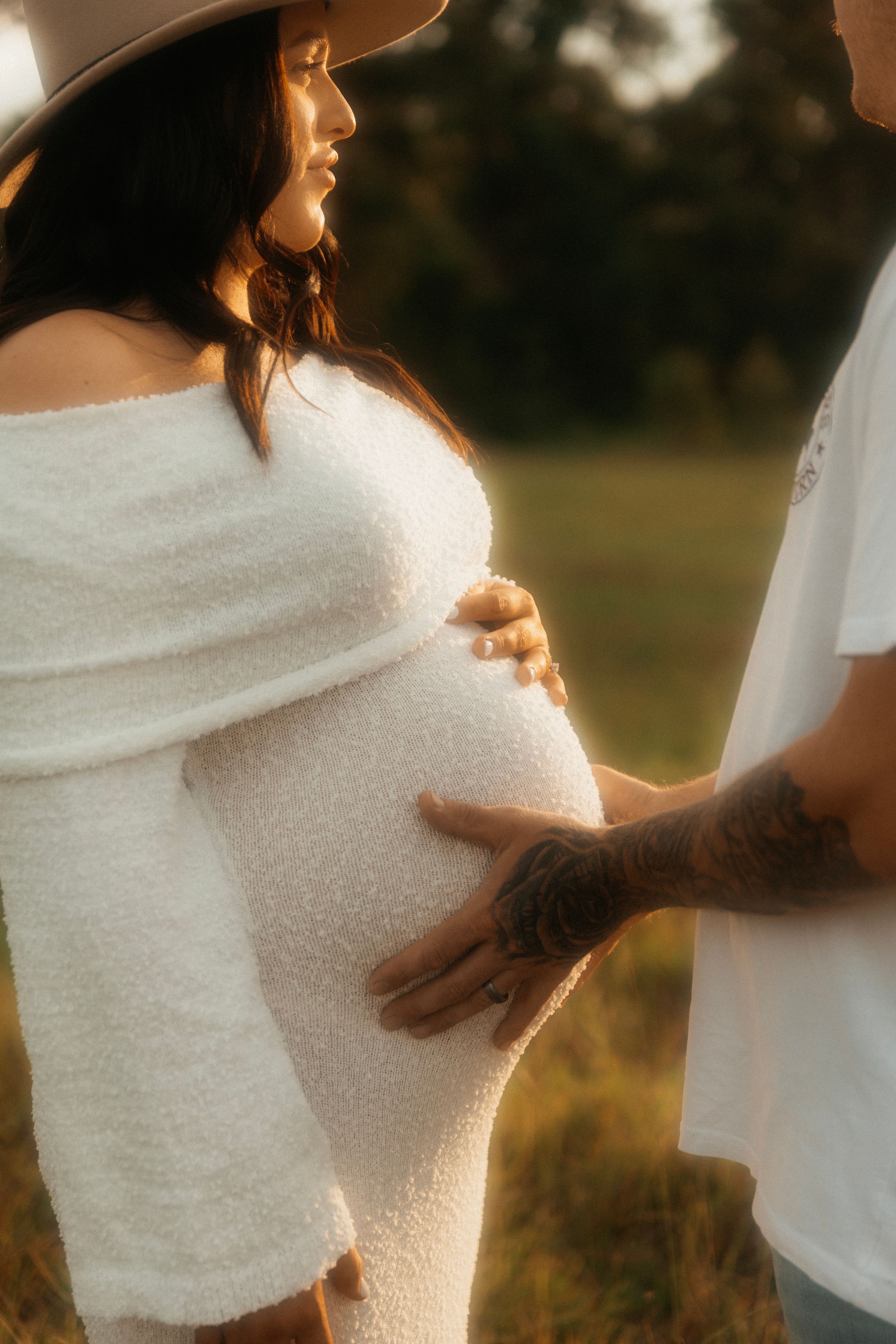 Close up of a gentle pregnancy photoshoot showing person in white dress with partner's hands on belly in warm sunlight.