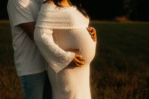A pregnant woman in a white lace dress stands in a golden field at sunset while being embraced from behind.