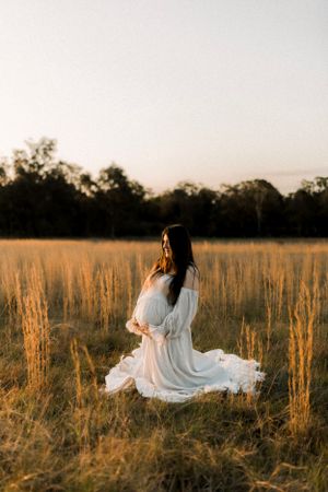 A woman in a flowing white dress sits gracefully in a golden field at sunset.