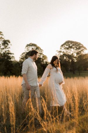 A couple walks hand in hand through tall golden grass at sunset.