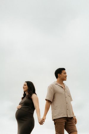 Couple holding hands against cloudy sky as they stand side by side in casual attire.