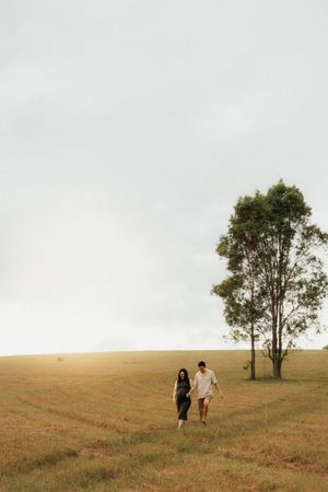 A couple walks together in a golden field with a lone tree against an overcast sky.
