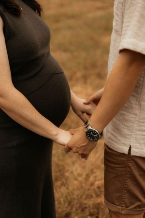Close-up of expecting couple holding hands, with a black maternity dress and wristwatch visible against a brown field.