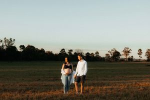 Expecting couple standing together in an open field at sunset with trees silhouetted in the background.