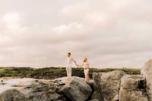 A couple stands together on coastal rocks during sunset.