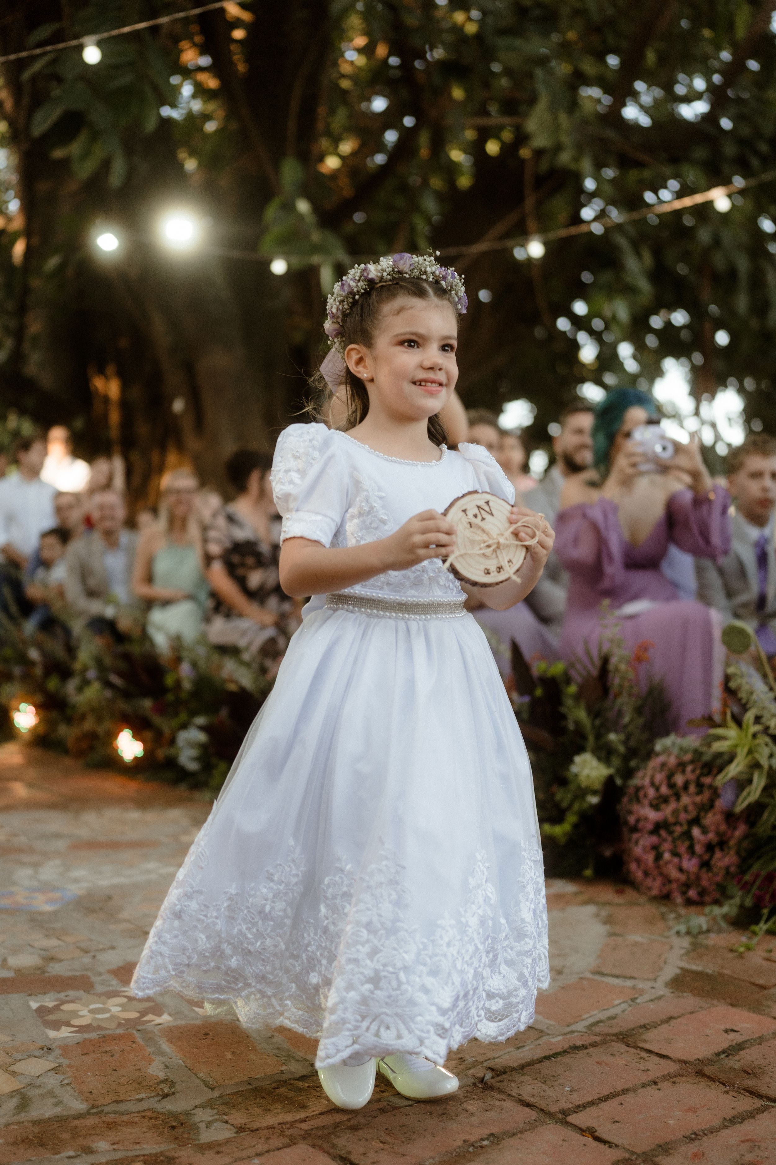 A young attendant in a white lace dress holds a decorative item during a wedding ceremony.