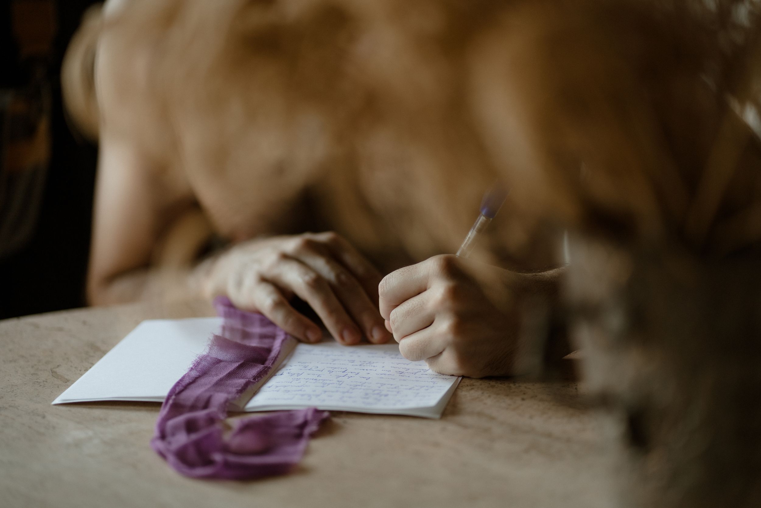 Hands writing on paper with a purple fabric accent in moody lighting.