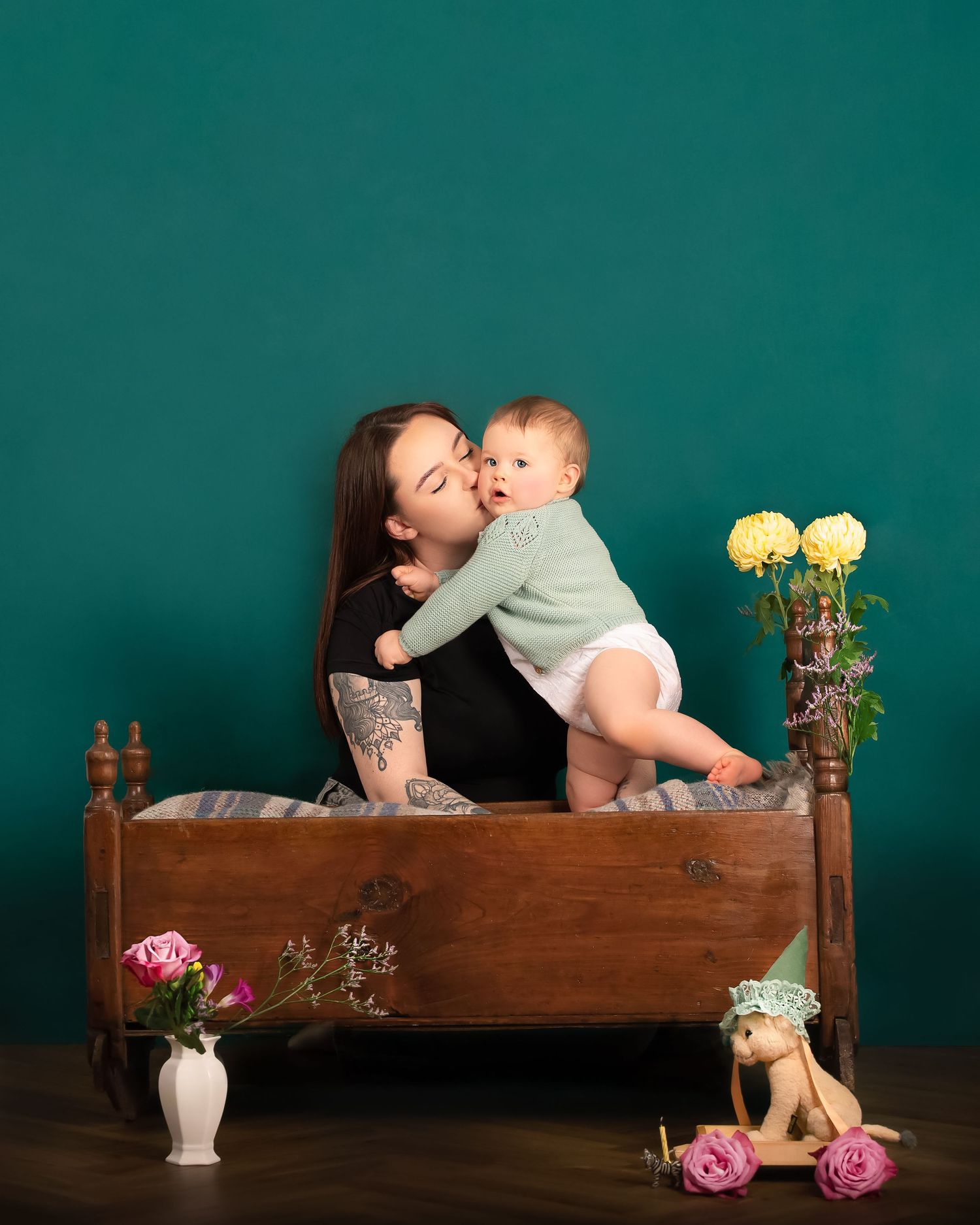 Sweet parent-child moment captured against a teal backdrop with antique wooden cradle and yellow flowers and white porcelain vase.