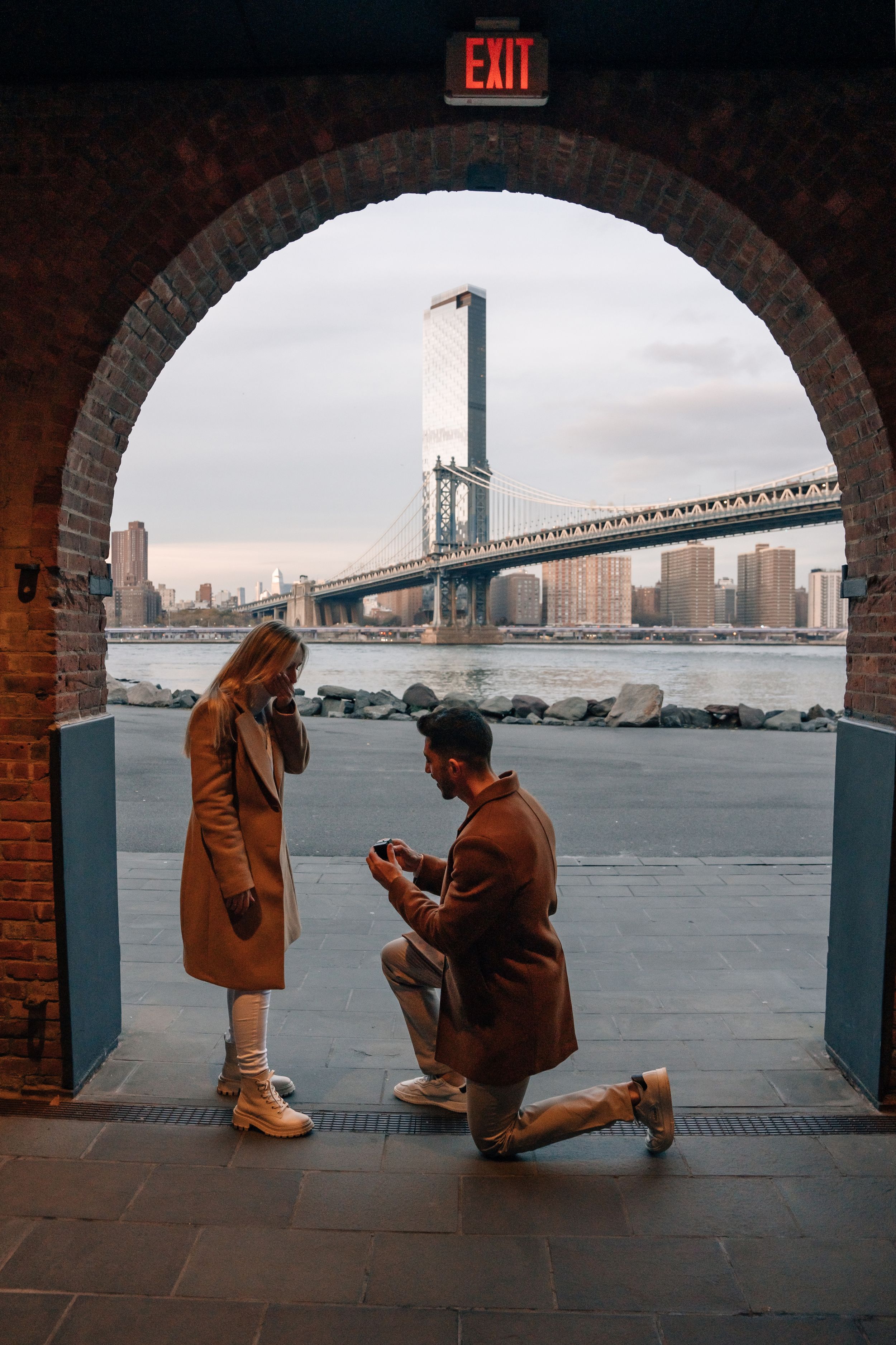 A romantic proposal moment framed by an archway with the Manhattan Bridge in the background.