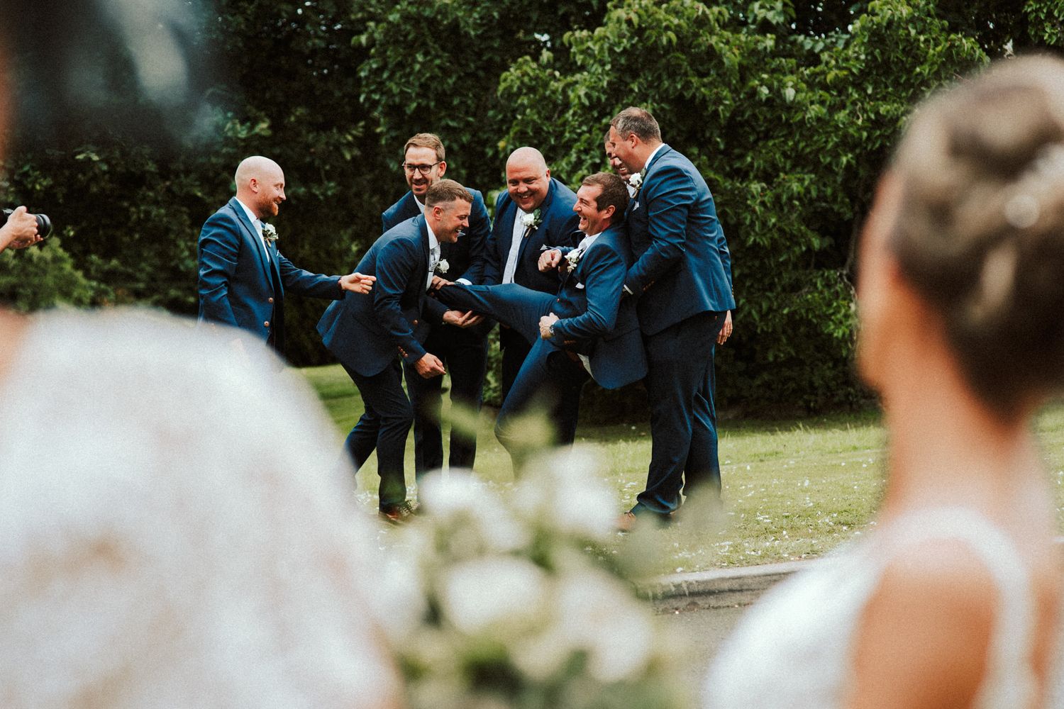 Groomsmen in blue suits jumping playfully for a fun wedding photo in an outdoor garden setting.